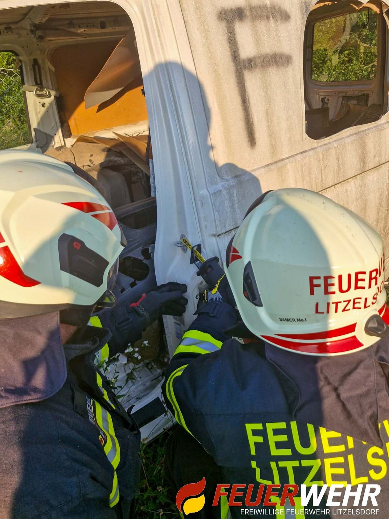 Zwei Feuerwehrleute in weißen Helmen arbeiten an einem beschädigten Fahrzeug, wahrscheinlich während einer Rettungsaktion. Einer hält ein Werkzeug, und beide tragen reflektierende Westen.
