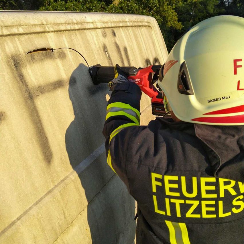 Ein Feuerwehrmann in Helm und Uniform mit den Worten 'Feuer Litzels' repariert eine Betonwand mit einem Elektrowerkzeug. Bäume und Schatten sind im Hintergrund sichtbar.