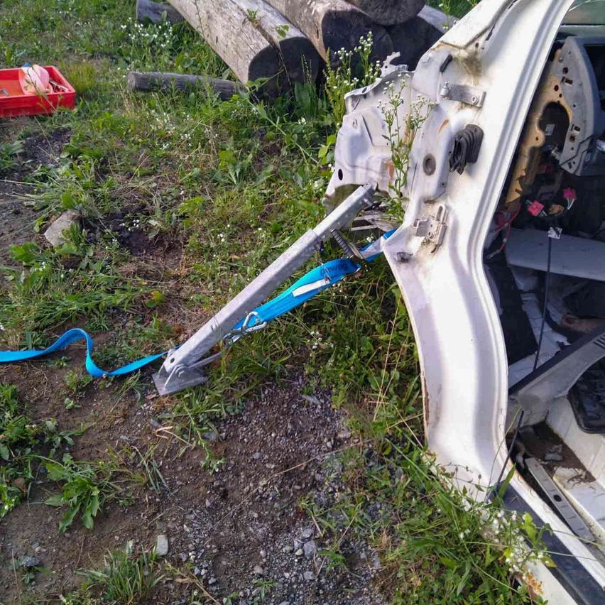 Eine beschädigte Autotür liegt mit einem blauen Seil auf Gras neben einem roten Werkzeugkasten und Holzstämmen.
