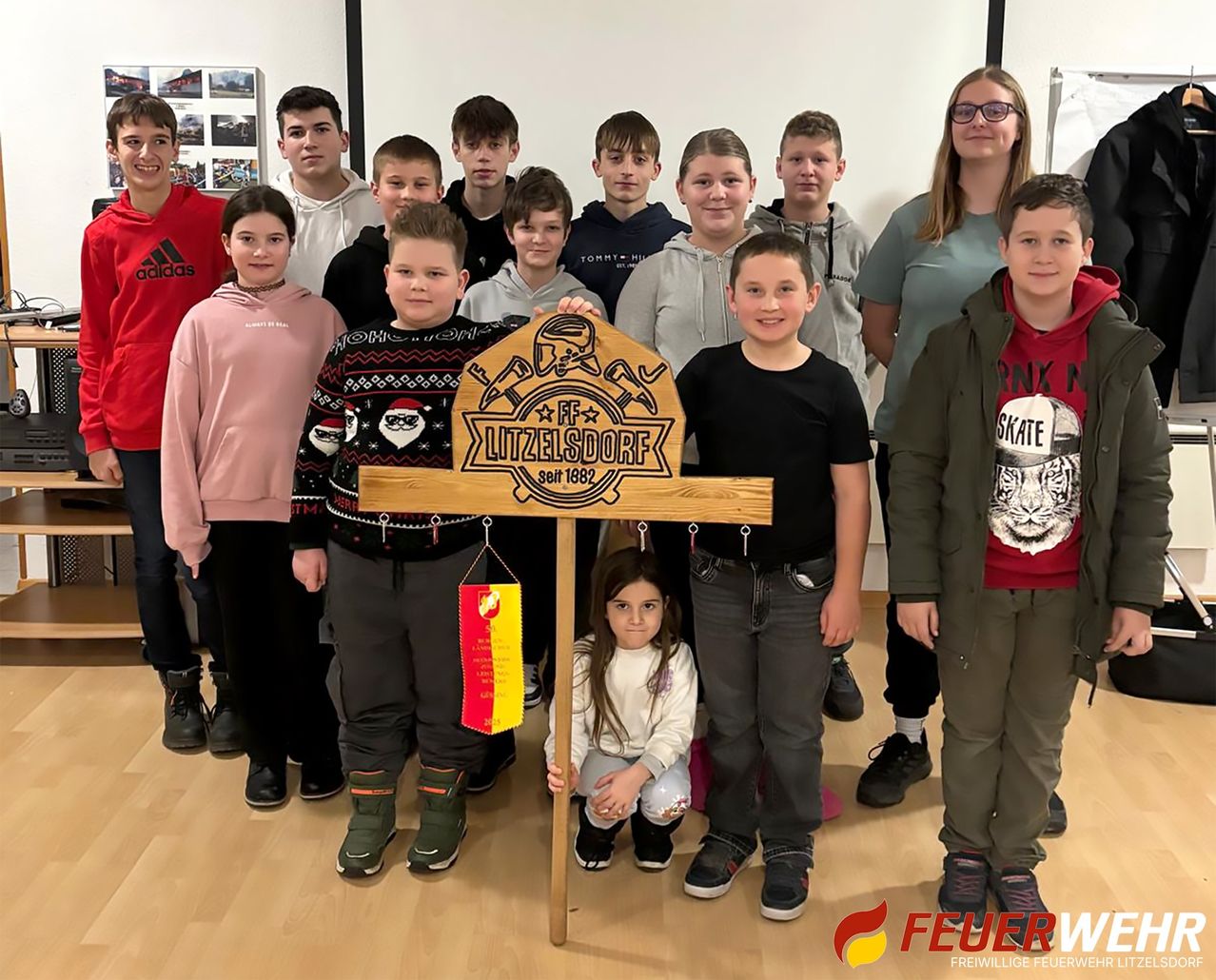 Eine Gruppe von Kindern und Erwachsenen posiert mit einem Holzschild für Litzelsdorf, an dem eine Fahne hängt. Sie stehen in einem Raum mit Holzboden.