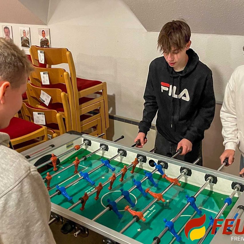 Zwei Jungen spielen Tischfußball in einem Raum mit einem Tischfußballtisch, während ein dritter Junge zuschaut. Der Raum hat weiße Wände, eine Decke und rot gepolsterte Stühle. Eine Person in einem weißen Shirt steht rechts.