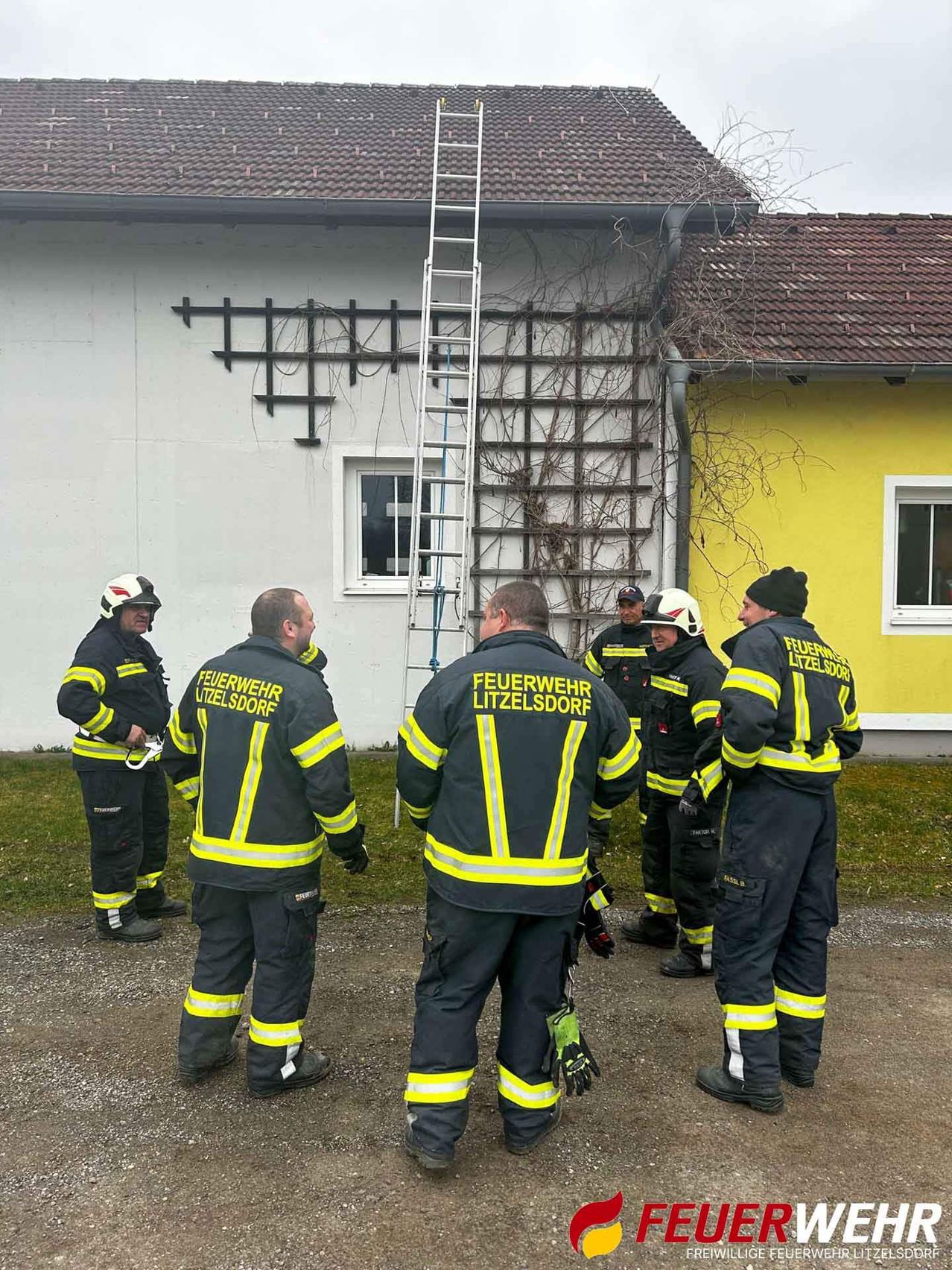 Eine Gruppe von Feuerwehrleuten in voller Uniform, einschließlich Helmen und Handschuhen, steht vor einem Gebäude mit einer Leiter und Weinranken.