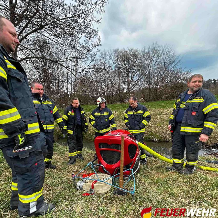 Eine Gruppe von Feuerwehrleuten steht um einen roten Feuerwehrwagen mit einer Schlauch. Sie befinden sich auf einer grasigen Fläche mit einem kleinen Bach im Hintergrund. Die Feuerwehrleute tragen Uniformen und Helme.
