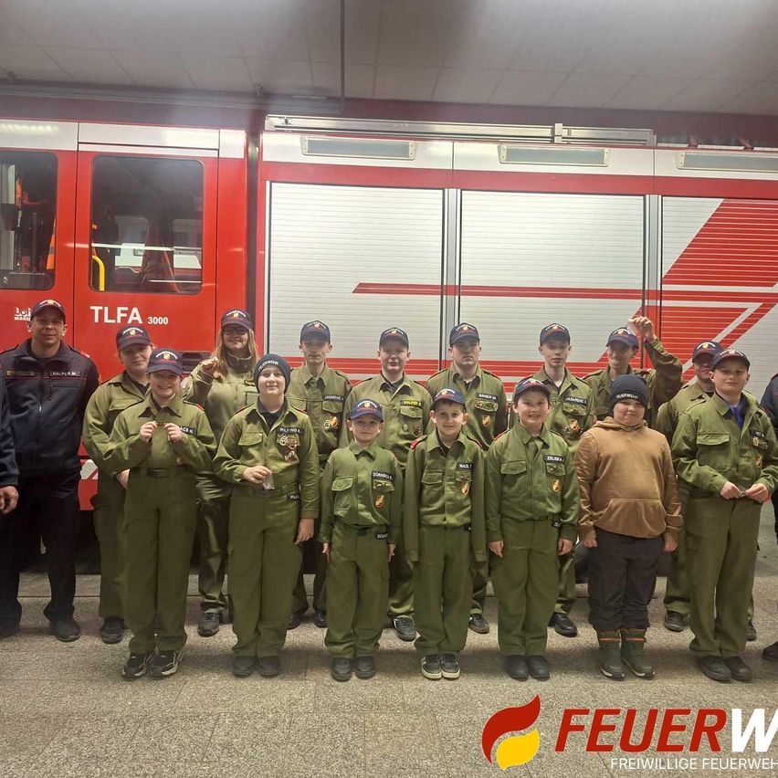 Eine Gruppe junger Menschen in grünen Uniformen, die vor einem Feuerwehrauto stehen, mit dem Wort 'Feuer' in der unteren rechten Ecke.