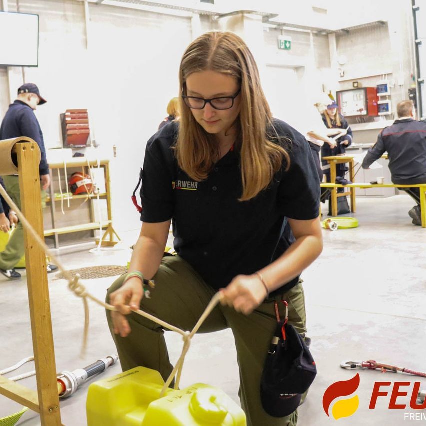 Eine Frau in einer Werkstatt übt das Knotenlegen an einem Seil. Sie trägt ein schwarzes Shirt mit Logo. Hinter ihr arbeitet eine weitere Person an einem Tisch. Verschiedene Werkzeuge und Ausrüstung sind umher verstreut.