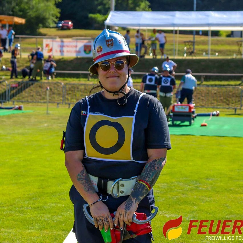 Eine Frau mit Helm und Sonnenbrille steht auf einem grünen Feld und hält einen Feuerlöscher. Im Hintergrund stehen Menschen unter einem Zelt, und andere sind auf dem Feld.