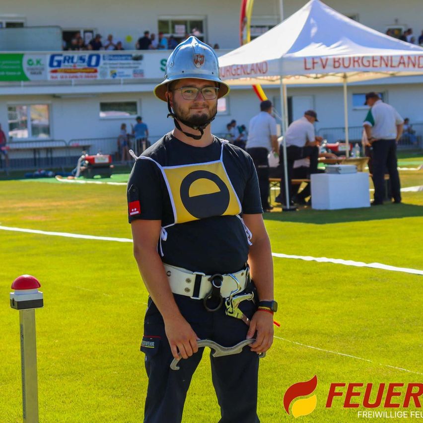 Ein Mann mit Helm und Gurt steht auf einem Feld mit einem Zelt und einem weißen Gebäude im Hintergrund.