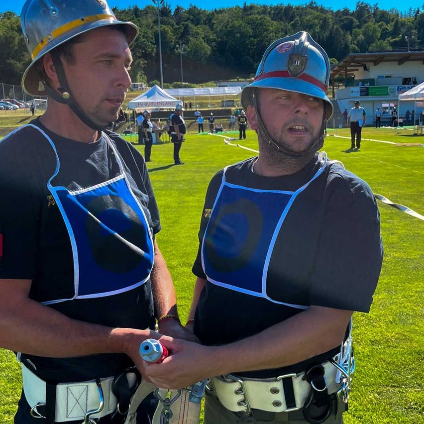 Zwei Männer in Feuerwehruniformen stehen auf einem Feld mit Bäumen im Hintergrund. Einer hält ein Werkzeug und beide tragen Helme. Menschen und Zelte sind in der Ferne.