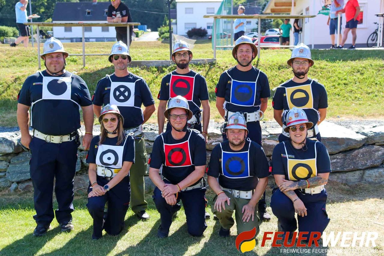 Eine Gruppe von Feuerwehrleuten in Uniformen mit Helmen und Weste, posiert für ein Foto auf einem Rasenfeld. Das Wort 'FEUER' ist auf der rechten unteren Seite gedruckt.