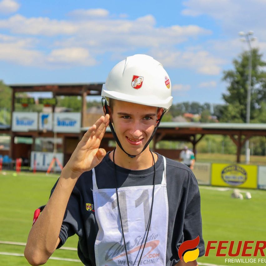 Ein junger Mann mit einem weißen Helm mit rotem Logo und der Nummer 11 auf seinem Shirt salutiert auf einem Rasenfeld mit verschwommenem Hintergrund.