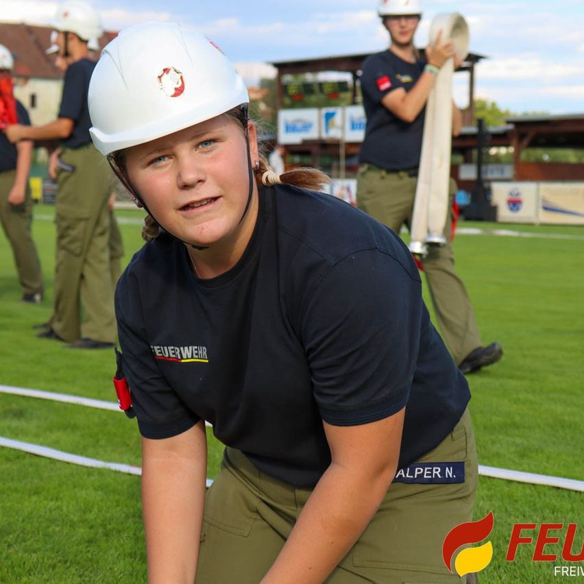 Eine junge Frau in einem weißen Helm und einem schwarzen Shirt hockt auf einem Feld mit anderen uniformierten Personen.