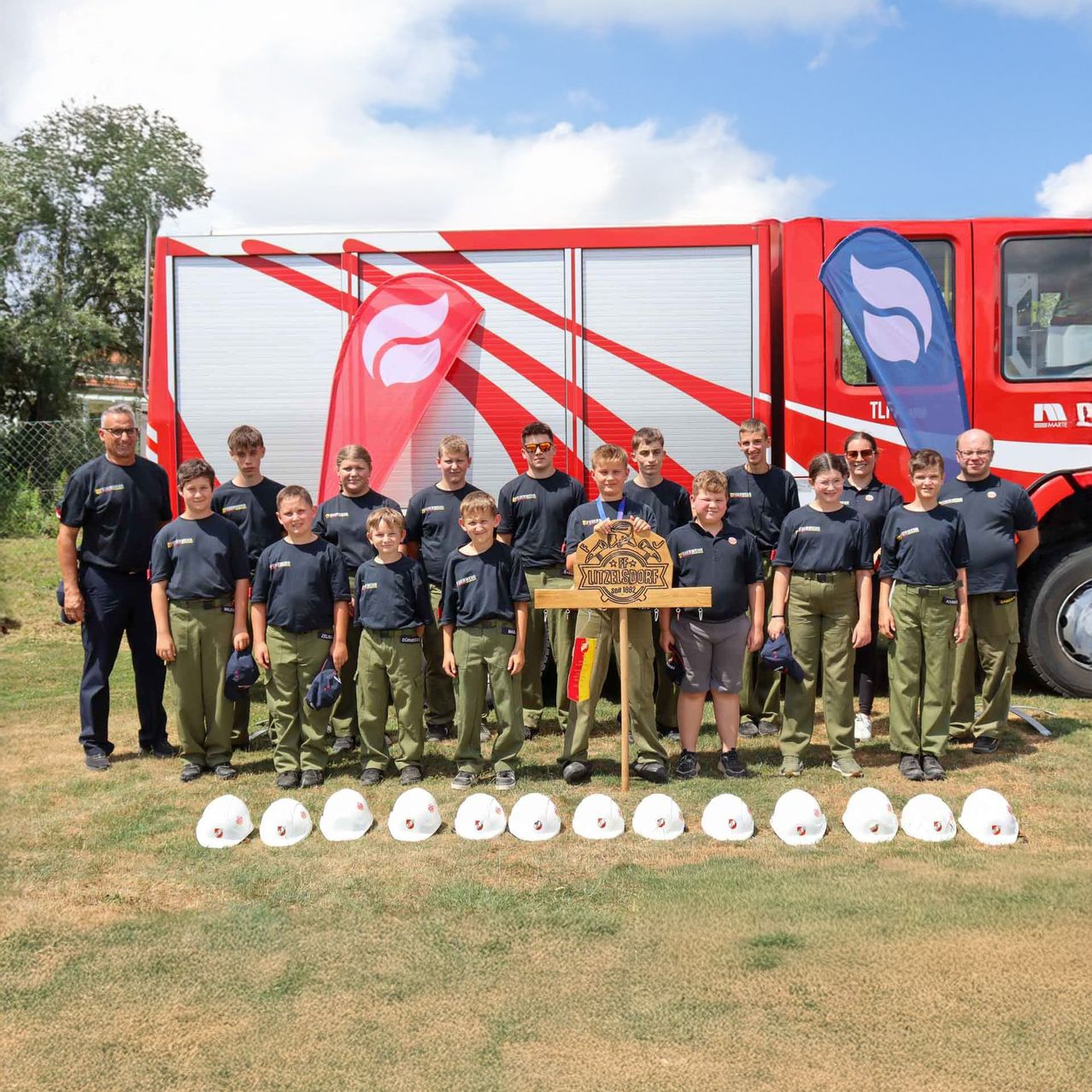 Eine Gruppe junger Menschen in Uniform vor einem Feuerwehrwagen, mit Helmen und einer Holztafel im Vordergrund.