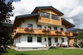 Ein zweistöckiges weißes Haus mit einem hölzernen Balkon, geschmückt mit Blumenkästen, in einem üppigen grünen Bereich mit Bergen im Hintergrund gelegen.