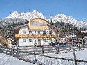 Ein großes weißes Chalet mit Holzakzenten befindet sich in einer verschneiten Landschaft mit einem Gebirgshintergrund. Das Gebäude verfügt über mehrere Fenster und Balkone. In der Nähe umgibt ein Holzzaun einen verschneiten Bereich.