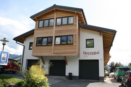 Ein zweistöckiges Chalet mit Holzfassade und drei Garagen, beschriftet mit Hoyergut, befindet sich in einer Bergregion mit üppiger Grünfläche und geparkten Autos.