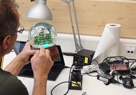 Ein Mann arbeitet an einer Schneekugel mit einer Weihnachtsbaum-Innenseite. Er sitzt an einem Schreibtisch mit Laptop und verschiedenen elektronischen Komponenten.