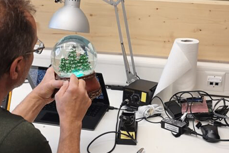 Ein Mann arbeitet an einer Schneekugel mit einer Weihnachtsbaum-Innenseite. Er sitzt an einem Schreibtisch mit Laptop und verschiedenen elektronischen Komponenten.