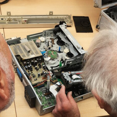 Ein älterer Mann repariert ein Gerät mit einer Leiterplatte auf einem Schreibtisch.