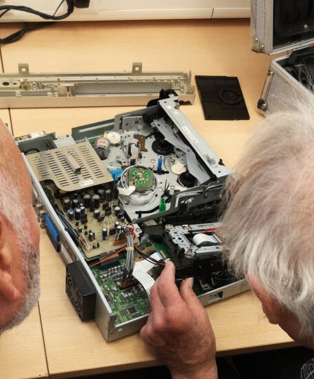 Ein älterer Mann repariert ein Gerät mit einer Leiterplatte auf einem Schreibtisch.