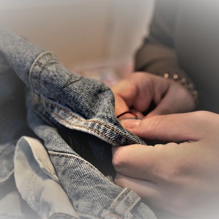 Ein Paar Jeans wird von jemandem repariert, wobei der Fokus auf dem Nähen liegt. Die Jeans sehen abgenutzt aus, werden aber sorgfältig repariert.