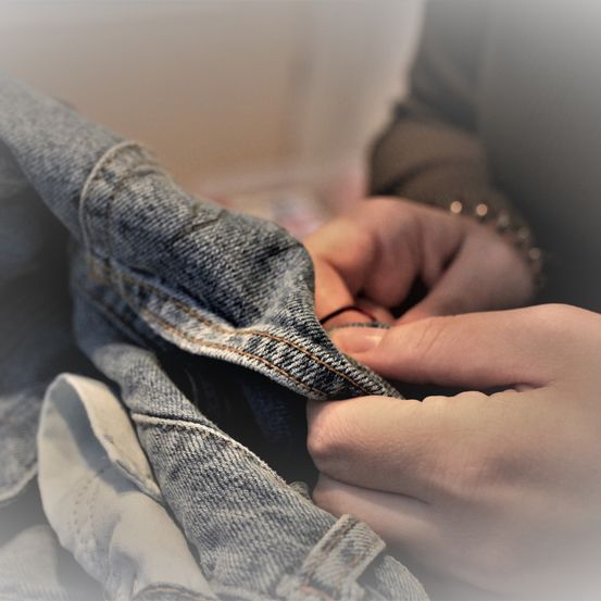Ein Paar Jeans wird von jemandem repariert, wobei der Fokus auf dem Nähen liegt. Die Jeans sehen abgenutzt aus, werden aber sorgfältig repariert.