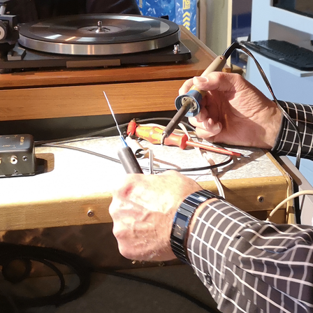 Eine Person benutzt einen Lötkolben auf einem Gerät mit Kabeln, in der Nähe eines Plattenspielers.