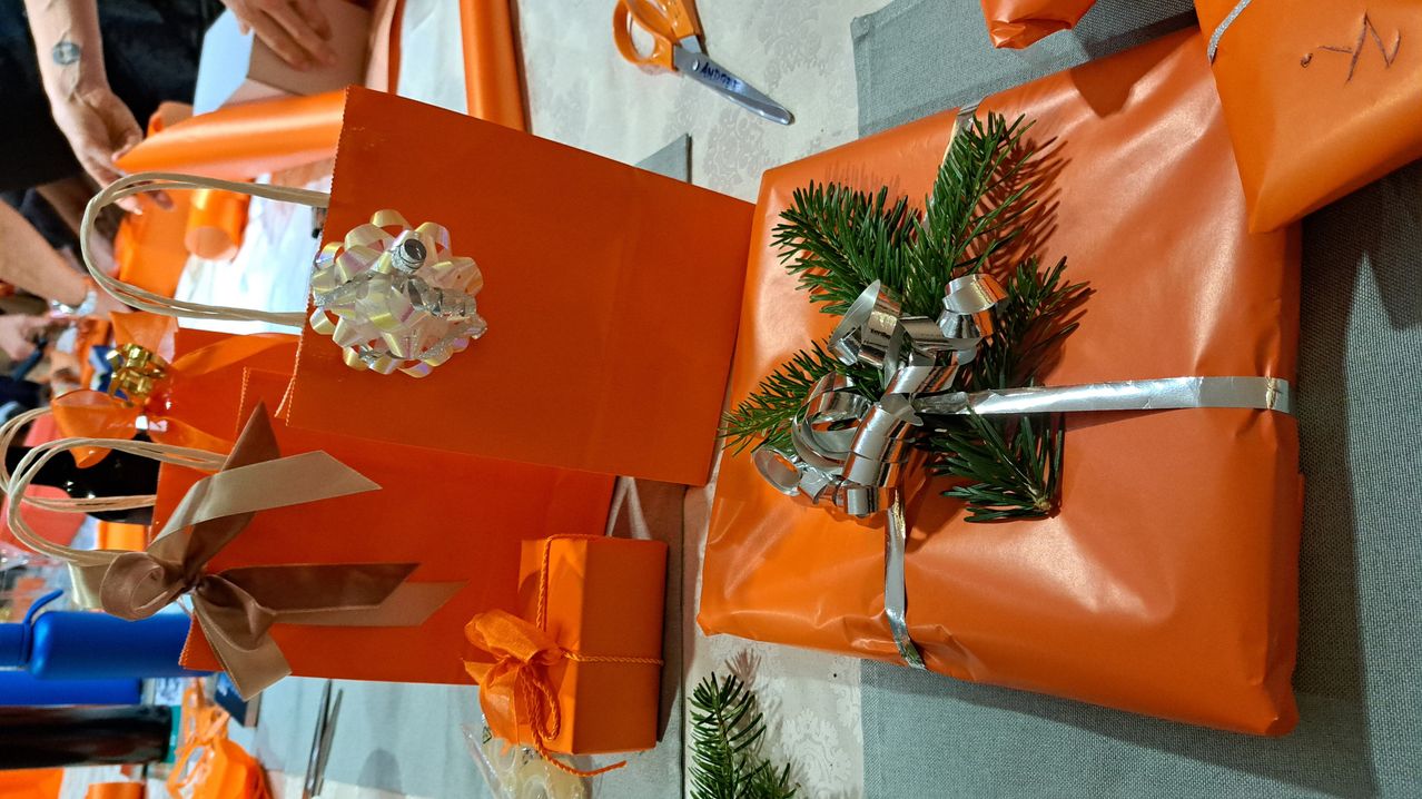 Mehrere orange verpackte Geschenke mit dekorativen Elementen, einschließlich einer Tannenzweig und einer glänzenden Schleife, liegen auf einem Tisch mit einer Schere und einem Lineal.