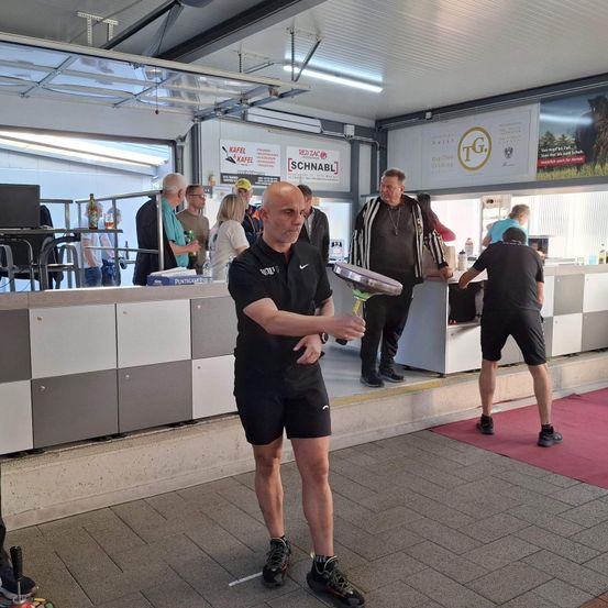 Eine Indoor-Sportveranstaltung, ein älterer Mann übt mit einem Schläger, während andere plaudern und ein Tresen mit Schildern sichtbar ist.