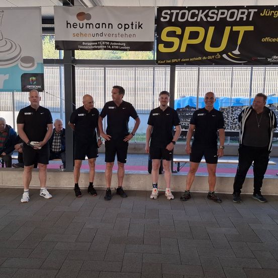 Eine Gruppe von Männern in Sportkleidung steht vor einem Gebäude mit Bannern, wobei zwei Banner sichtbar sind. Hinter der Gruppe sitzen Menschen.