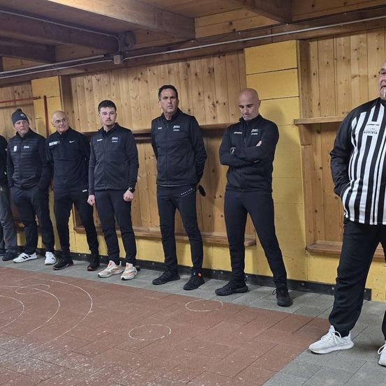 Sieben Männer in identischen schwarzen Jacken stehen in einer Reihe. Zwei haben die Arme verschränkt. Hinter ihnen steht ein Mann in einem schwarz-weiß gestreiften Hemd in der Nähe einer Holzwand.