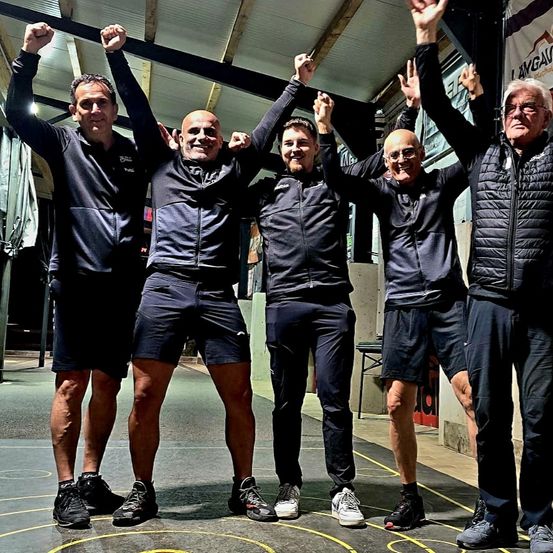 Fünf Männer stehen zusammen und heben alle ihre Arme zum Sieg. Sie sind in dunkler blauer Sportbekleidung gekleidet und feiern wahrscheinlich einen Sieg. Im Hintergrund befindet sich ein Gebäude mit einem Schild, möglicherweise ein Fitnessstudio.