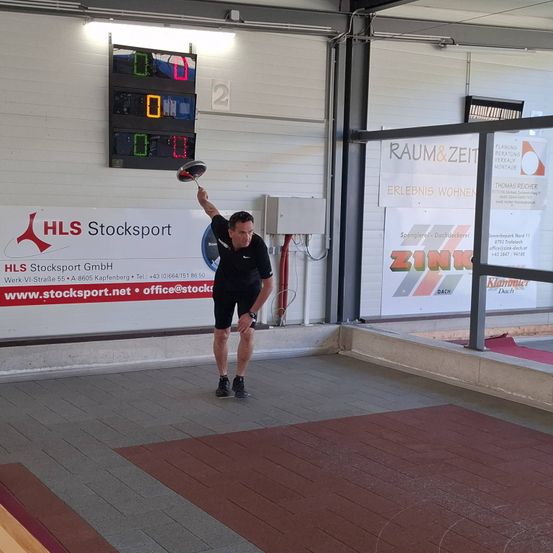 Ein Mann in schwarzem Shirt und Shorts wirft einen Frisbee in einer Sportanlage mit digitalem Punkteanzeiger.