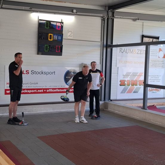 Drei Männer spielen drinnen eine Partie Boccia. Eine digitale Anzeigetafel zeigt die Punktzahl. Ein Mann steht, ein anderer bereitet sich zum Werfen vor und ein dritter beobachtet.