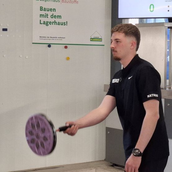Ein Mann hält einen Schläger mit einem violetten Scheiben und schwingt ihn, wahrscheinlich spielt er Badminton. Er trägt ein schwarzes Shirt mit 'RIZE' und 'MATHIAS' darauf. Hinter ihm ist eine Wand mit einem grünen Schild, das 'Bauen mit dem Lagerhaus' sagt.