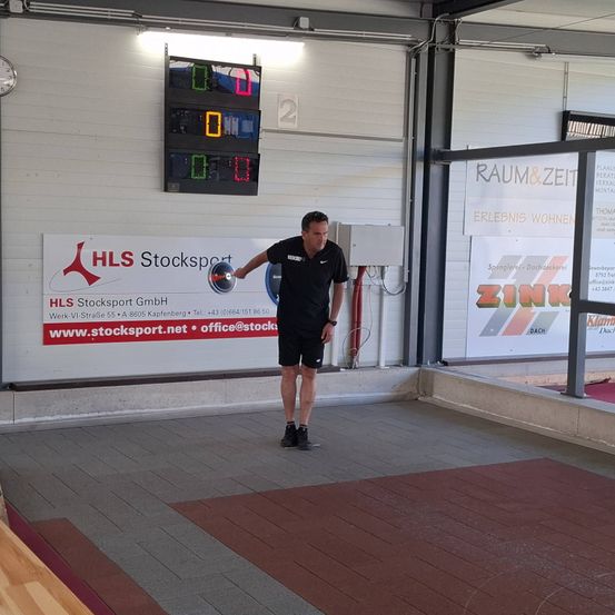 Ein Mann in einem schwarzen Shirt und Shorts wirft einen Diskus in einer Turnhalle mit einer Uhr an der Wand.