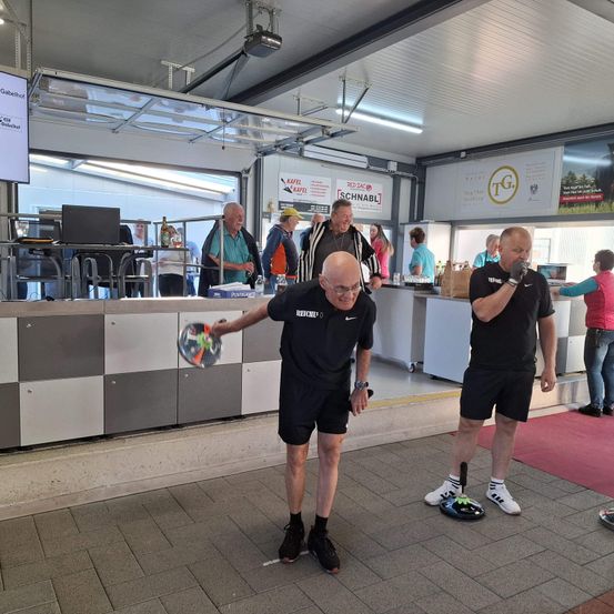 Zwei Männer in einem Saal spielen mit Frisbees. Sie sind in schwarzer Sportbekleidung gekleidet und scheinen Spaß zu haben. Hinter ihnen steht eine Theke mit einem Monitor und anderen Leuten.