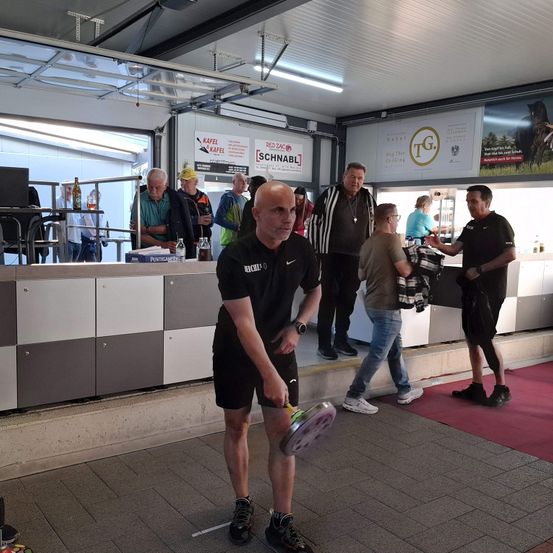 Ein Mann spielt Frisbee in einem Innenbereich. Mehrere Personen stehen herum. Der Bereich hat einen gefliesten Boden und einen roten Teppich. Ein Mann trägt ein schwarzes Shirt und Shorts.