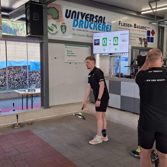 Zwei Männer spielen Tennis in einer Einrichtung mit einem Universal Druckerei Schild. Die Anzeigetafel zeigt 0-0. Ein Mann schlägt den Ball, während der andere ein Foto macht.