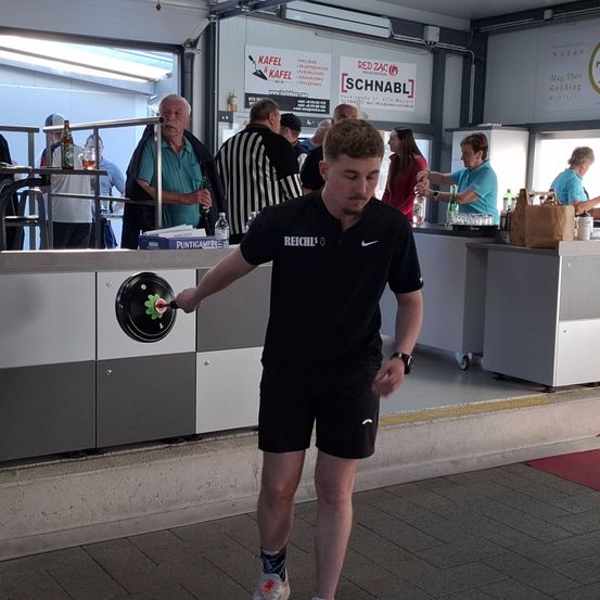 Ein junger Mann wirft einen Disc im Discgolf-Bereich, während Zuschauer zuschauen. Hinter ihm hat eine Theke Flaschen und einen braunen Papierbeutel. An der Wand befinden sich Werbungen für 'RED ZAC SCHNABEL' und 'KAFEL & KAFEL'.