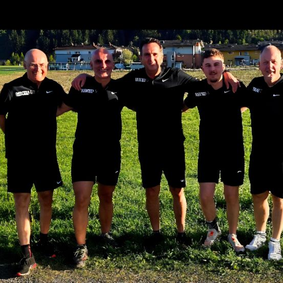 Fünf Männer in identischer schwarzer Sportbekleidung posieren für ein Foto auf einem Rasenfeld mit Häusern im Hintergrund.