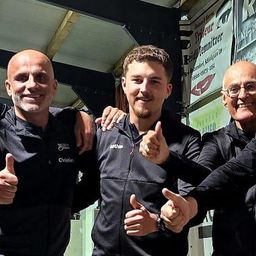 Fünf Männer in schwarzer Sportbekleidung stehen zusammen und posieren für ein Foto. Sie lächeln und geben Daumen hoch. Der Ort scheint eine Sporteinrichtung zu sein.
