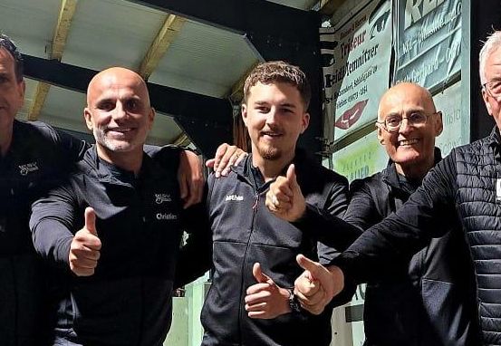 Fünf Männer in schwarzer Sportbekleidung stehen zusammen und posieren für ein Foto. Sie lächeln und geben Daumen hoch. Der Ort scheint eine Sporteinrichtung zu sein.