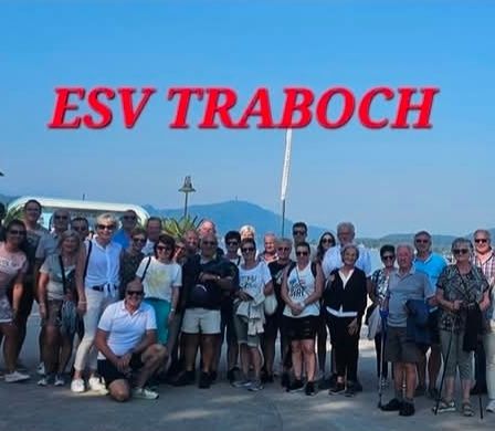 Eine große Gruppe von Menschen posiert für ein Foto am ESVT Traboch, vor einem See mit Bergen und einer Stadt im Hintergrund stehend.