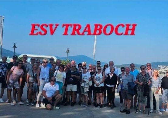Eine große Gruppe von Menschen posiert für ein Foto am ESVT Traboch, vor einem See mit Bergen und einer Stadt im Hintergrund stehend.