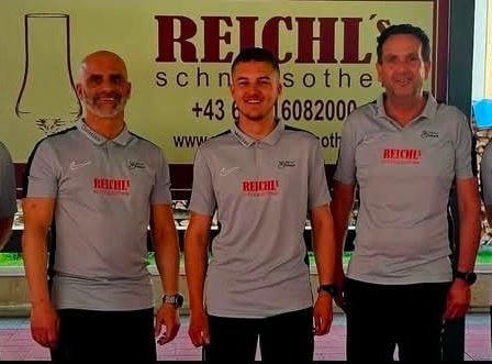 Fünf Männer in gleichen grauen Shirts stehen zusammen draußen und lächeln für die Kamera. Sie tragen gleiche schwarze Hosen. Das Banner hinter ihnen sagt 'REICHLS' und hat Kontaktdaten.