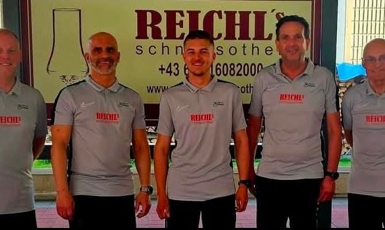 Fünf Männer in gleichen grauen Shirts stehen zusammen draußen und lächeln für die Kamera. Sie tragen gleiche schwarze Hosen. Das Banner hinter ihnen sagt 'REICHLS' und hat Kontaktdaten.
