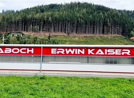 Ein großes Gebäude mit einem roten Schild, das 'ESV Traboch Erwin Kaiser Halle' besagt, gelegen an einem Hang mit üppigem Grün und Bäumen im Hintergrund.