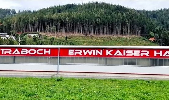 Ein großes Gebäude mit einem roten Schild, das 'ESV Traboch Erwin Kaiser Halle' besagt, gelegen an einem Hang mit üppigem Grün und Bäumen im Hintergrund.