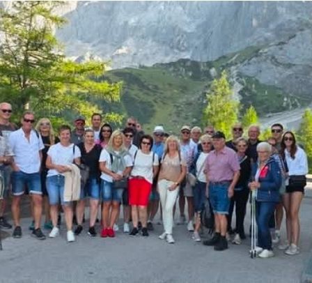 Eine Gruppe von Menschen steht vor einer Bergkette, lächelt und posiert für ein Foto. Sie sind in lässiger Sommerkleidung gekleidet.