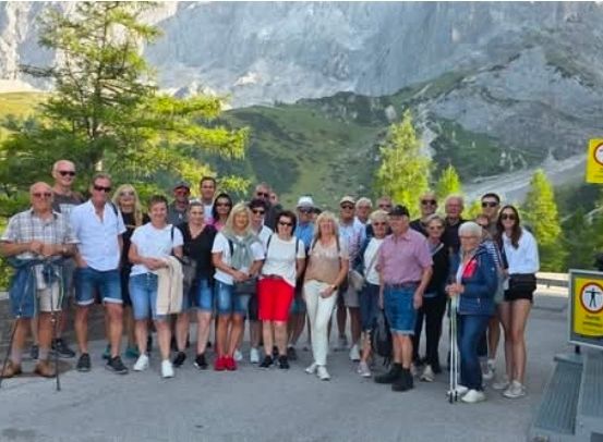 Eine Gruppe von Menschen steht vor einer Bergkette, lächelt und posiert für ein Foto. Sie sind in lässiger Sommerkleidung gekleidet.